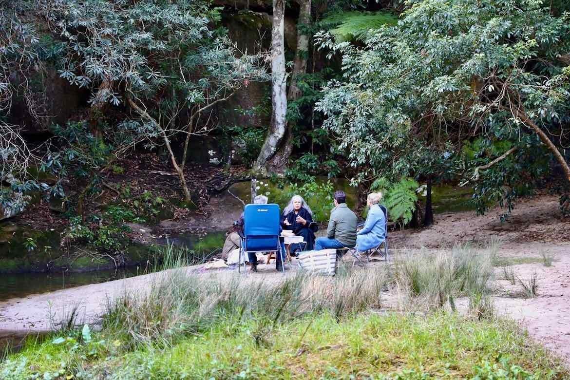 A group of landscape architects and Susan Moylan-Coombs sit in the bush on blue camp chairs.