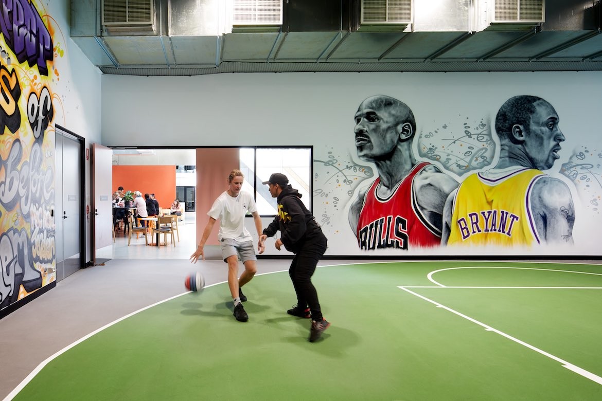 Two people play basketball on an indoor court surrounded by murals in Hester Hornbrook Academy by Gray Puksand.