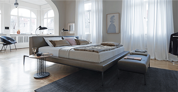 Jaan Bed, designed by EOOS. The lightness of being. Not bright and loud. The striking thing about the Jaan bed is its charmingly elegant allure. The designers made the upholstered bed float: generous lounging spaces on a delicate framework – seemingly weightless, Jaan shapes the room. Supremely soft and coolly casual. Poetry in the form of clean lines. Supple leather or natural fabrics form the headboard and upholstered framework. Additional comfort is provided by the matt shimmering servers and the board in black glass or striking saddle leather. High-class details, elaborately and carefully crafted. A wonderful place for all those essentials – your favourite book, your watch, a reading lamp. As obvious as it is special. 