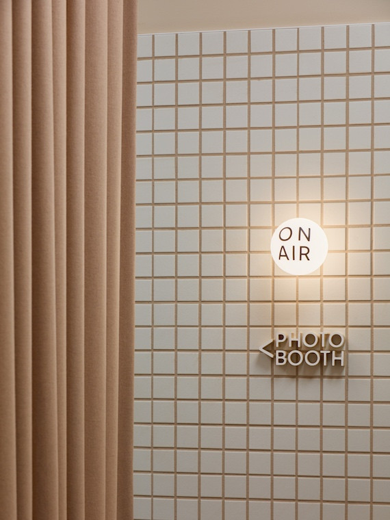 Cream tiles with dusty pink grout cover one wall that has a sign pointing to the photo booth on it in New Me medical centre by Rezen Studio.