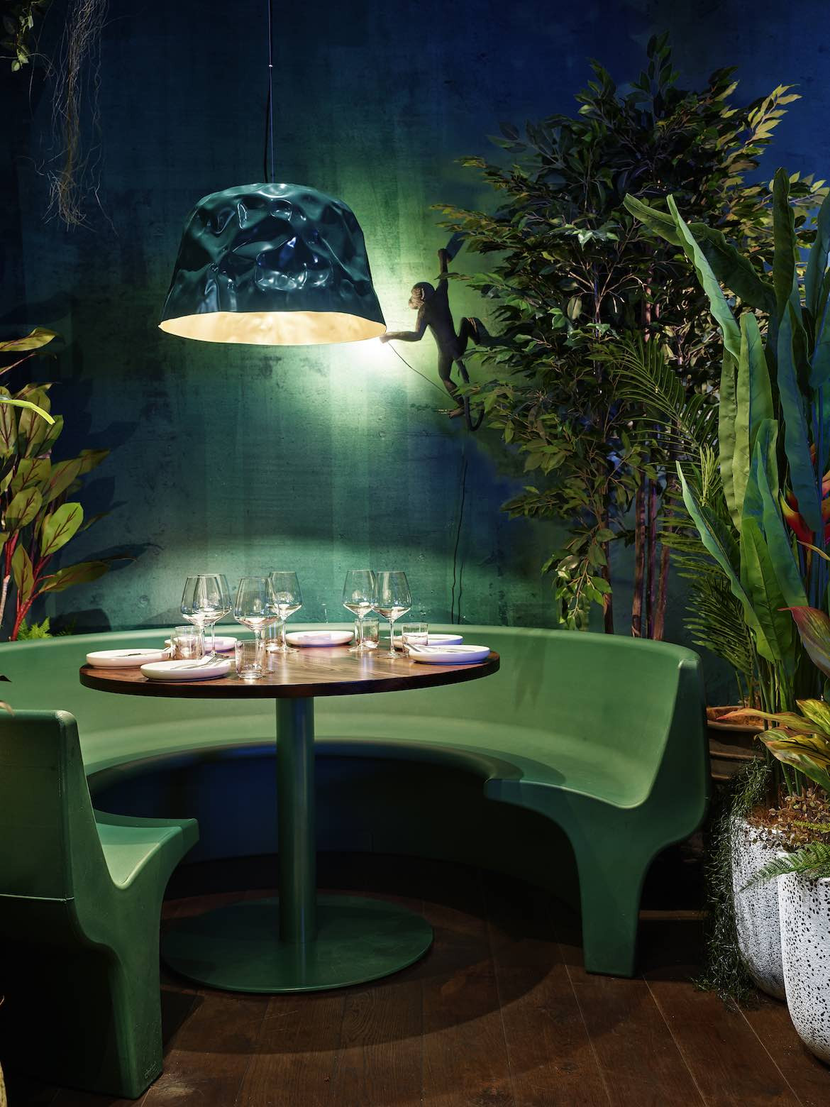 A green derlot booth seat underneath a hanging pendant light and a monkey, surrounded by plants.