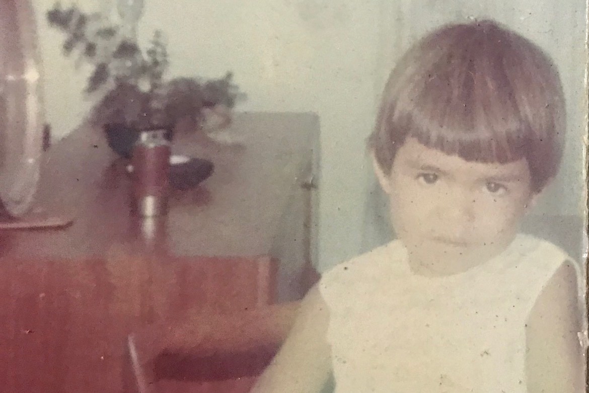 Susan Moylan-Coombs at age 3 with straight brown hair wears a white dress and looks at the camera.