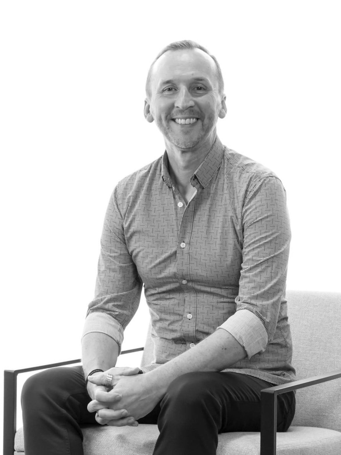 A black and white photo of Mark Freeman from Gray Puksand sitting on an armchair with his hands clasped, smiling at the camera.