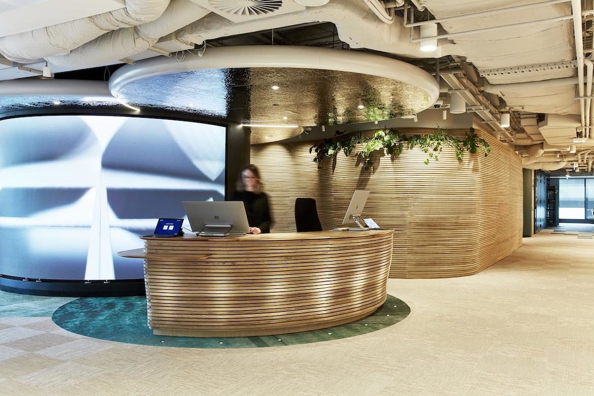 A blurry woman stands behind a curved, wood pannelled desk in front of a blue and white illuminated graphic screen at the Microsoft Sydney headquarters designed by group GSA.