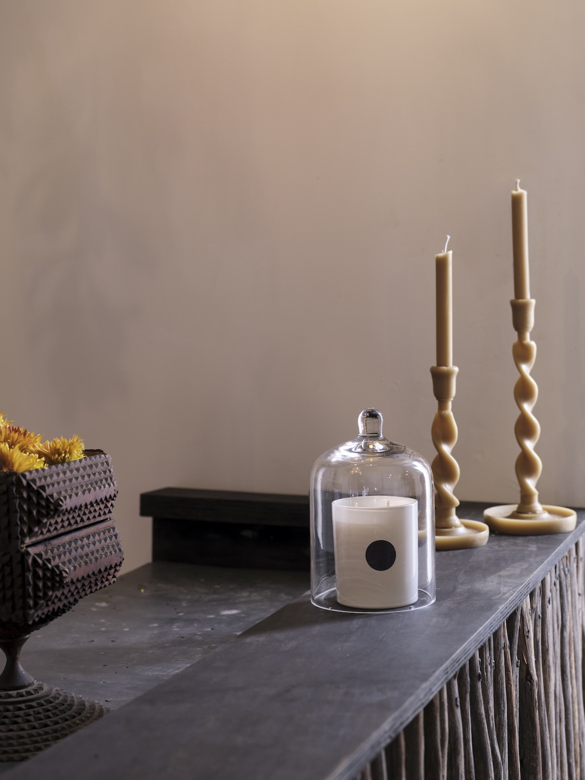 Two twirling beeswax candles on a wooden counter, next to a scented candle by The Raconteur