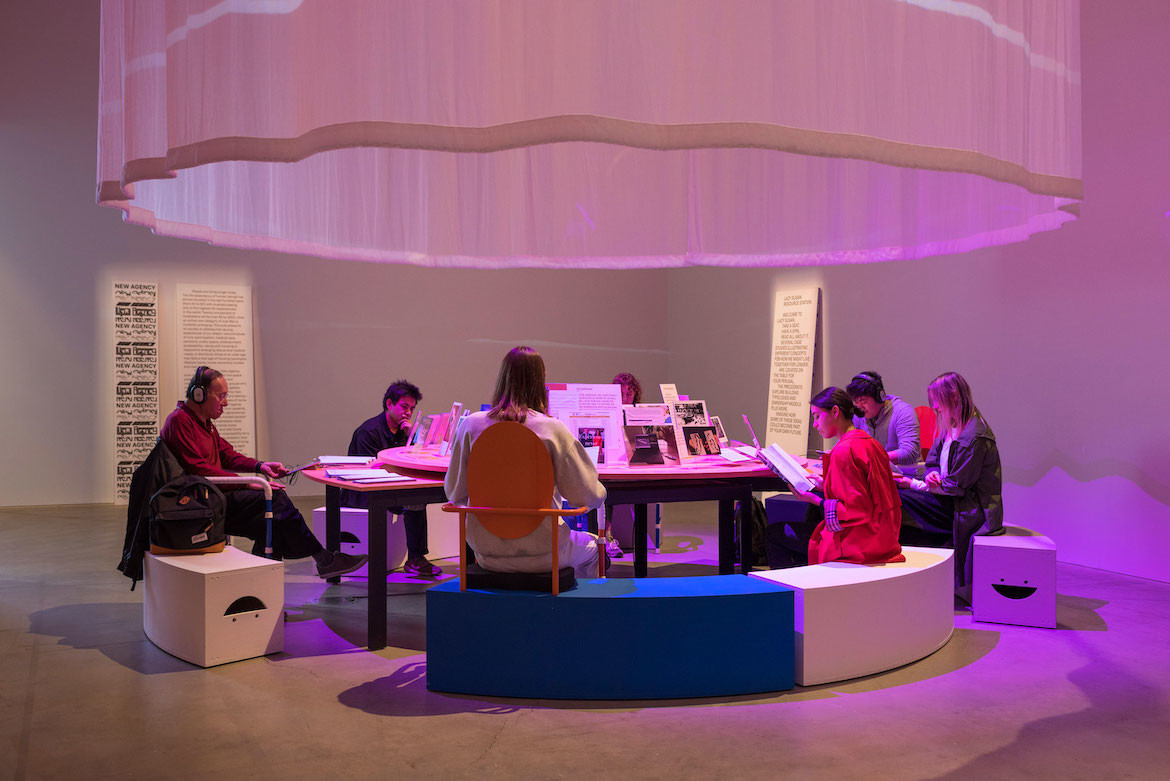 People sit around a reading table on blue and white bench seats in the Sibling exhibition reading information about New Agency and shrouded in purple light.