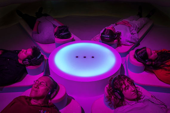 Five people lie on beds, shrouded in purple light, listening to an installation at the New Agency exhibition.