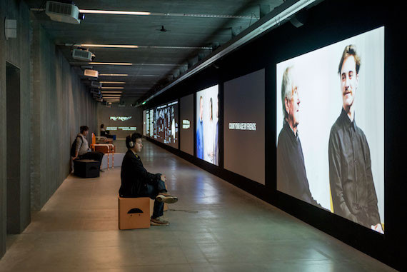 A long corridor has projector screens running down it, displaying videos and text at the New Agency exhibition.