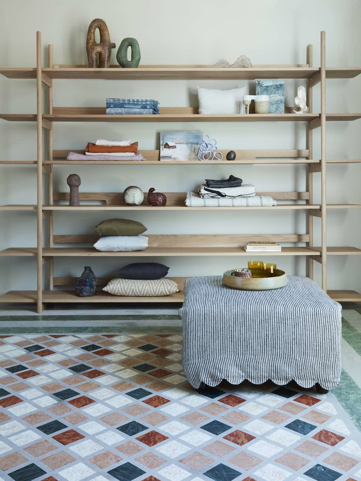 A pinstripe linen fabric by Cultiver drapes an ottoman next to wooden shelving and on top of the colourful handpainted floor.