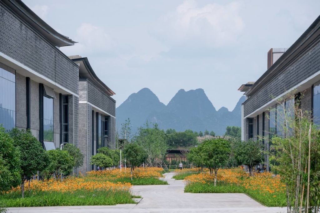 Between two integral buildings and patches of green grass with orange flowers, the peaks of mountains are visible in the distance.