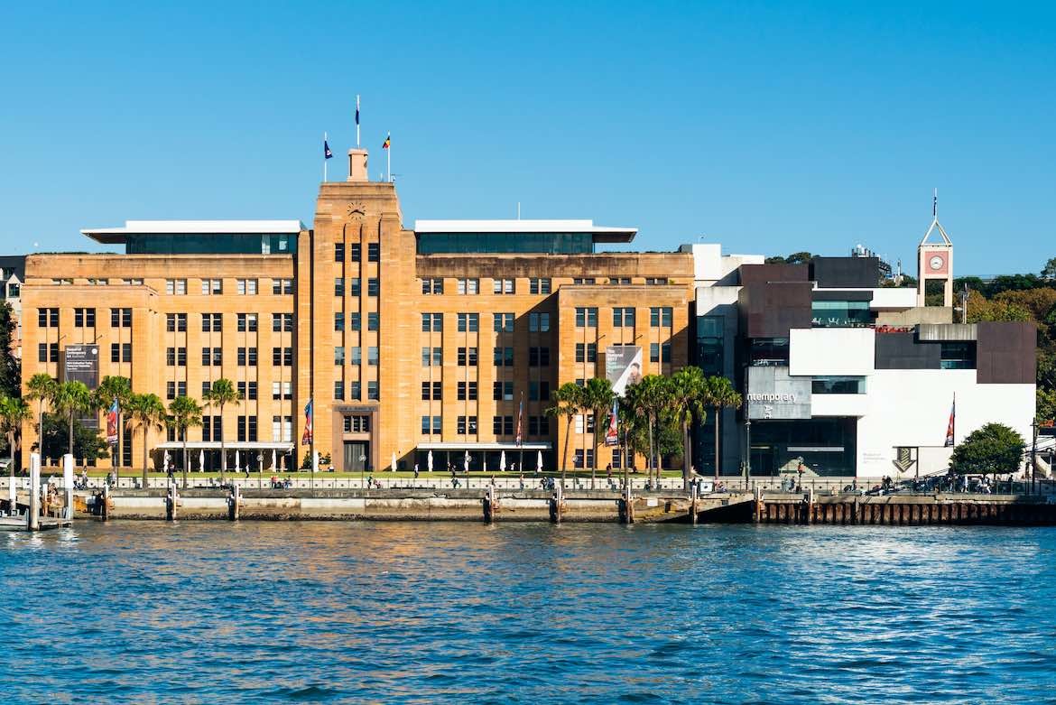 A view across water of the MCA – an historic sandstone building next to a contemporary brown and white building – from Australian Architecture: A History.