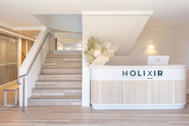 A curving white and cane reception desk that has Holixir written in teal letters across it, by Building Guild.