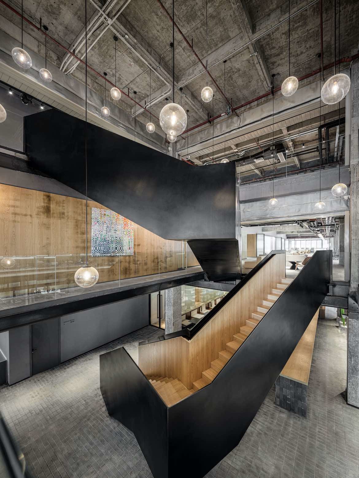 A wooden staircase in the centre of the office is painted black on its exterior, under an exposed ceiling and hanging pendant lights in the Yeahka office by JSPA Design.